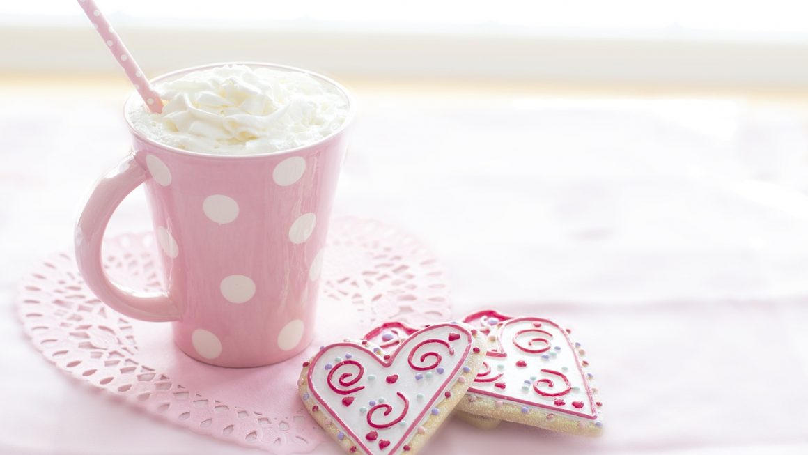 Un mug rose à pois blancs rempli de boisson chaude garnie de crème fouettée et une paille rose, accompagné de biscuits en forme de cœur décorés pour la Saint-Valentin, posés sur un napperon en dentelle rose, évoquant une douce célébration de la Saint-Valentin célibataire.