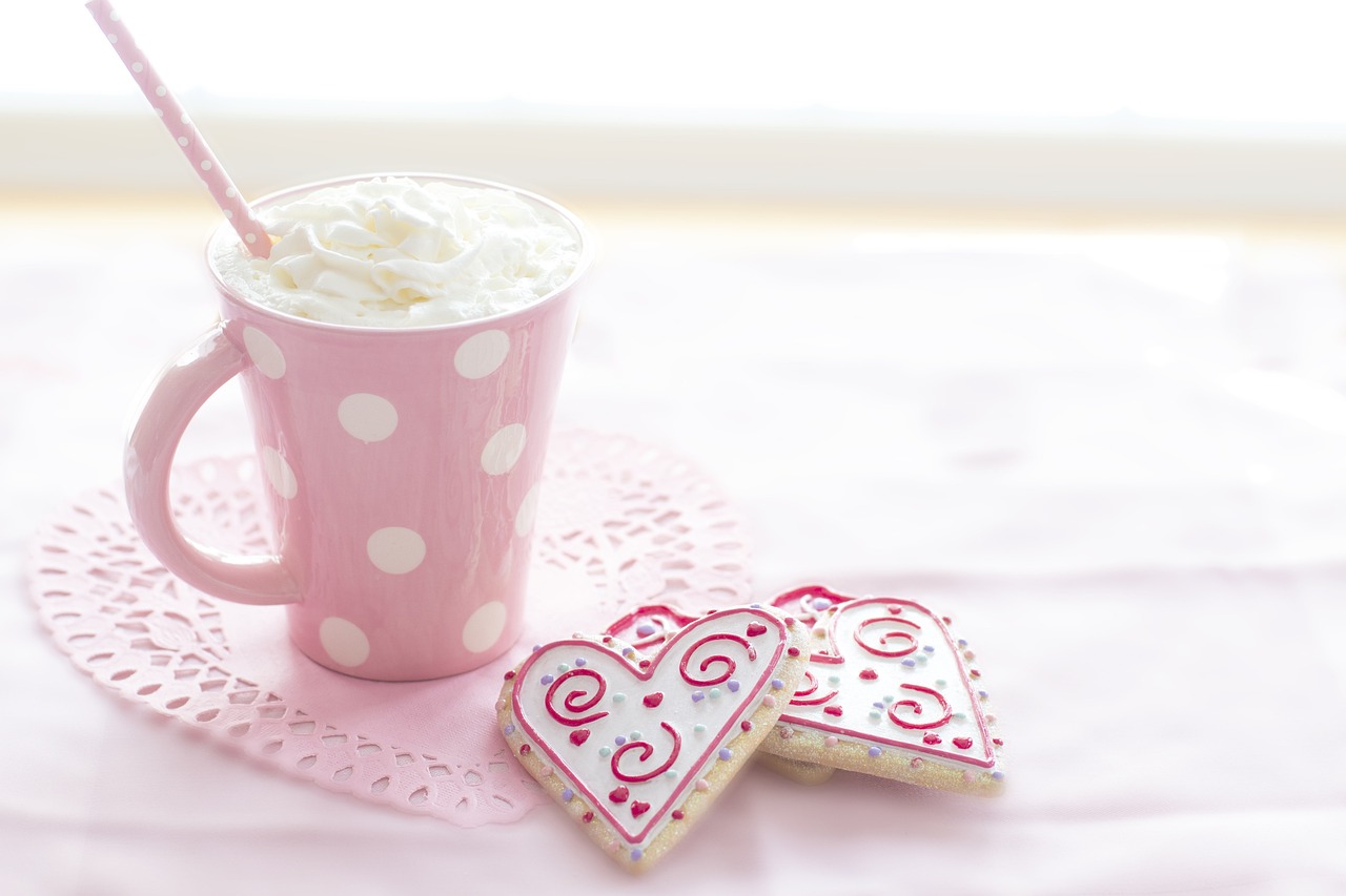 Un mug rose à pois blancs rempli de boisson chaude garnie de crème fouettée et une paille rose, accompagné de biscuits en forme de cœur décorés pour la Saint-Valentin, posés sur un napperon en dentelle rose, évoquant une douce célébration de la Saint-Valentin célibataire.