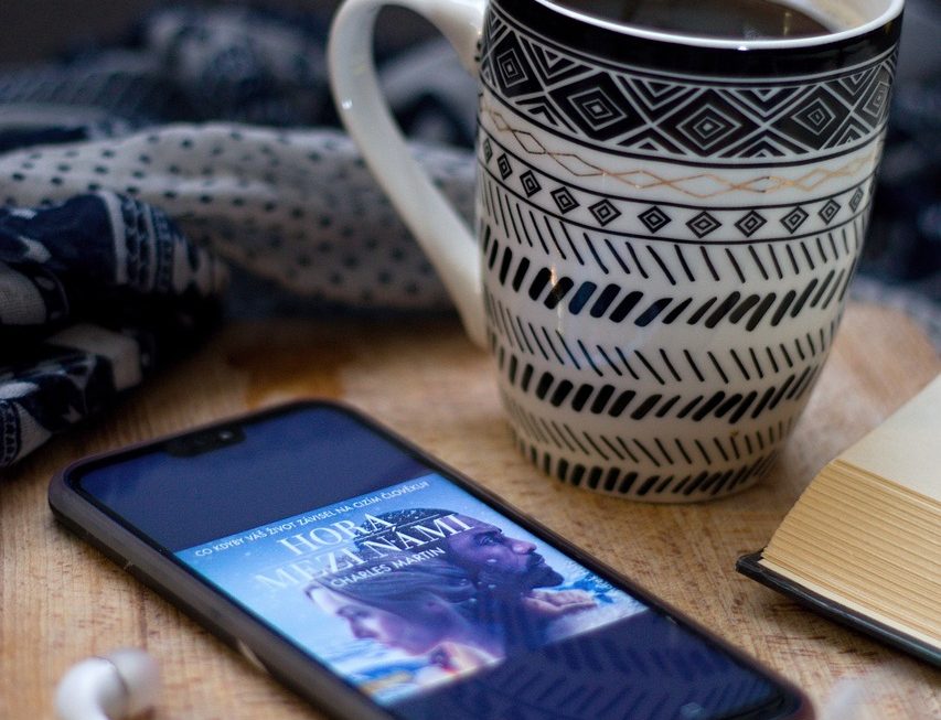 Une tasse de café décorée avec des motifs noirs et blancs est posée sur une table en bois, à côté d'un téléphone affichant la couverture d'un ebook intitulé "Hora Mezatami" de Charles Maxin. Des écouteurs blancs sont branchés au téléphone. Comment la technologie transforme notre manière de lire ?
