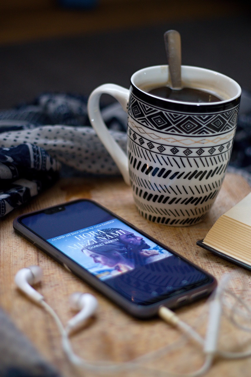 Une tasse de café décorée avec des motifs noirs et blancs est posée sur une table en bois, à côté d'un téléphone affichant la couverture d'un ebook intitulé "Hora Mezatami" de Charles Maxin. Des écouteurs blancs sont branchés au téléphone. Comment la technologie transforme notre manière de lire ?