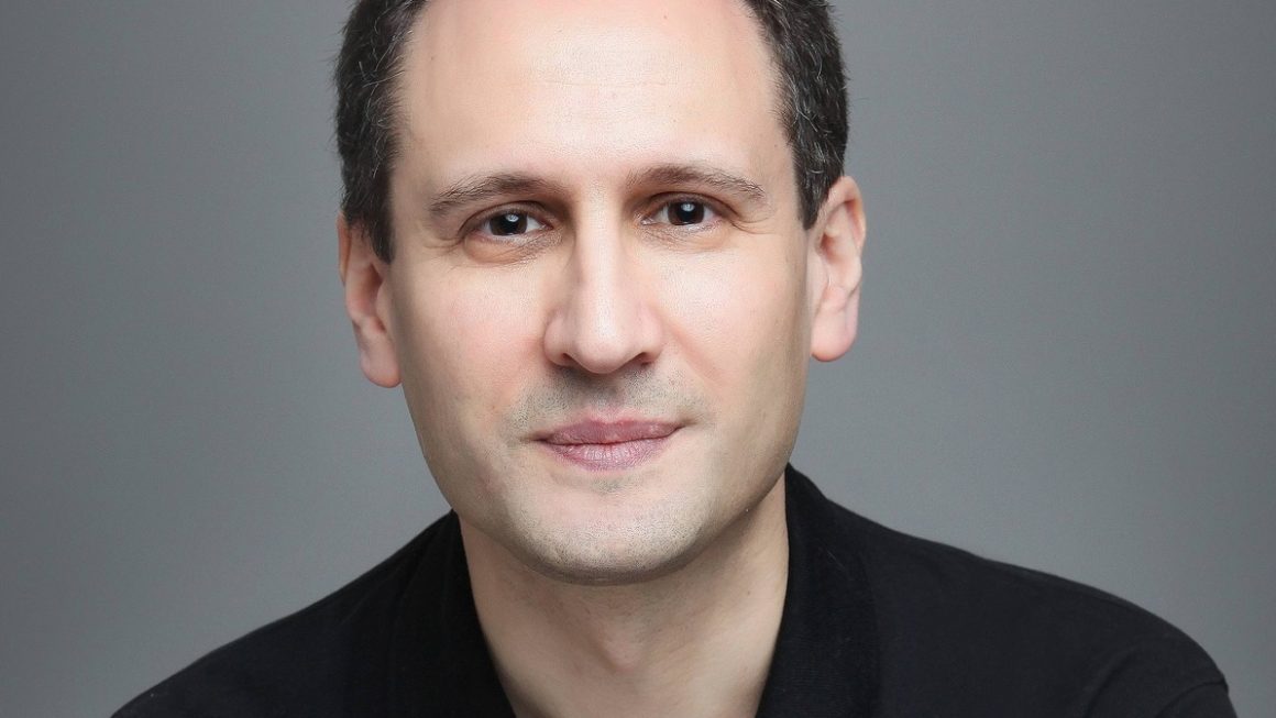 Portrait de William Alexandre, auteur du livre Perdre son job, encaisser et rebondir, vêtu d’un polo noir, les bras croisés et arborant une montre élégante. Il pose avec un léger sourire, regardant directement l’objectif