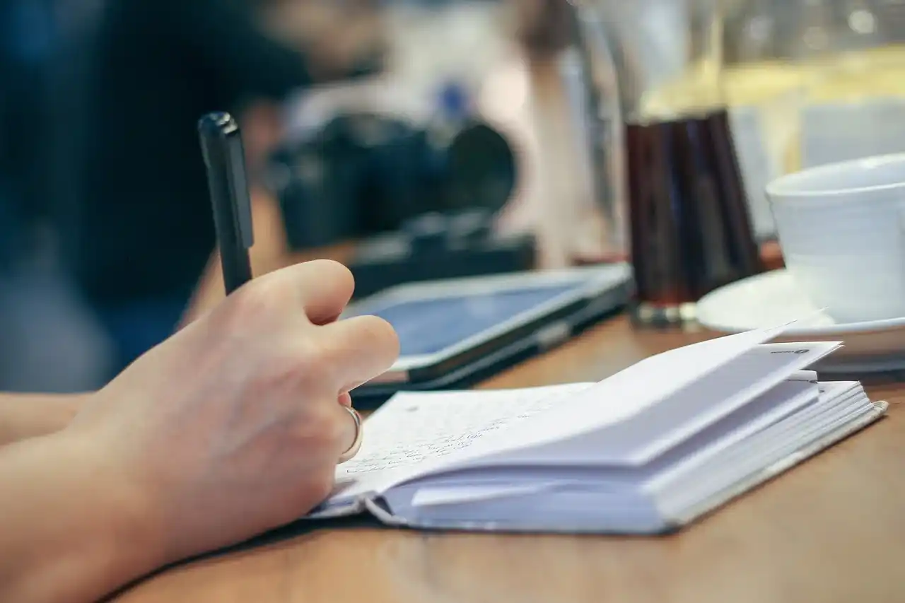 Une main féminine avec une bague écrit dans un carnet à spirale sur une table en bois. L’atmosphère est calme et studieuse, avec un ordinateur fermé, une tasse de café et un verre d’eau en arrière-plan flou. La scène évoque la concentration et le travail créatif, peut-être lié à l’écriture ou au brainstorming pour éviter les erreurs dès les premières pages d'un roman.