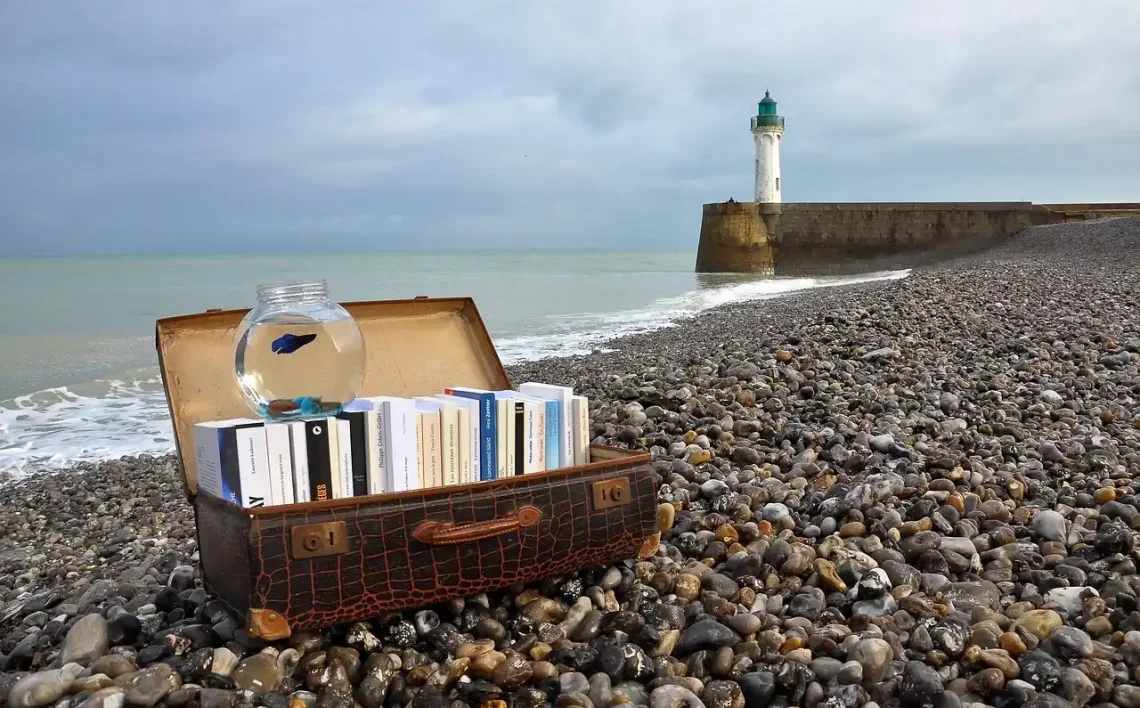 Valise ouverte remplie de livres posée sur une plage de galets, avec un phare en arrière-plan et un bocal contenant un poisson ; illustration symbolique d’une chronique littéraire professionnelle invitant au voyage et à la découverte d’auteurs.